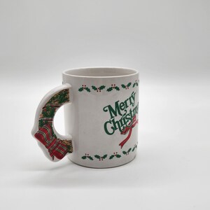 Puede incluir: Taza de cer&aacute;mica blanca con un dise&ntilde;o de acebo verde y bayas rojas alrededor del borde. La taza tiene un asa 3D con forma de corona de Navidad con cinta roja y verde. La taza dice "Merry Christmas" en letras verdes.