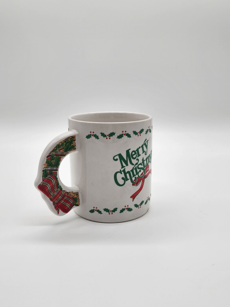 Puede incluir: Taza de cer&aacute;mica blanca con un dise&ntilde;o de acebo verde y bayas rojas alrededor del borde. La taza tiene un asa 3D con forma de corona de Navidad con cinta roja y verde. La taza dice "Merry Christmas" en letras verdes.