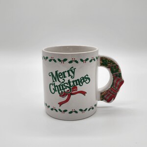 Puede incluir: Taza de cer&aacute;mica blanca con un borde de acebo verde y el texto "Merry Christmas" en letras verdes con una cinta roja. El asa tiene forma de calcet&iacute;n de Navidad con rayas rojas y verdes.