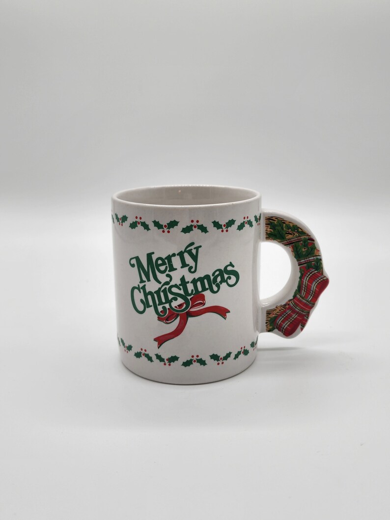 Puede incluir: Taza de cer&aacute;mica blanca con un borde de acebo verde y el texto "Merry Christmas" en letras verdes con una cinta roja. El asa tiene forma de calcet&iacute;n de Navidad con rayas rojas y verdes.