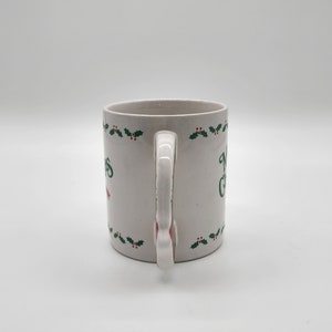 Puede incluir: Taza de cer&aacute;mica blanca con un dise&ntilde;o de hojas de acebo verdes y el texto "Feliz Navidad".