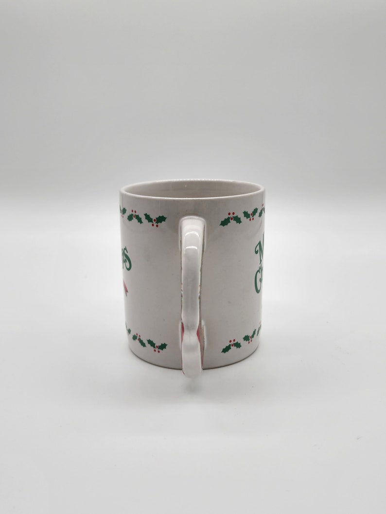 Puede incluir: Taza de cer&aacute;mica blanca con un dise&ntilde;o de hojas de acebo verdes y el texto "Feliz Navidad".