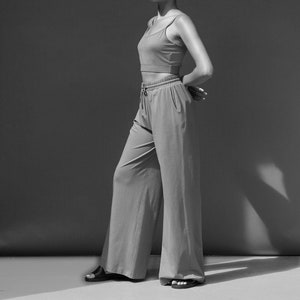 May include: A woman wearing a black and white outfit. She is wearing a black tank top and white wide-leg trousers. She is standing in front of a grey background.
