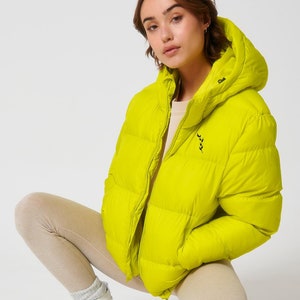 May include: A bright yellow puffer jacket with a hood and a small leaf logo on the left chest. The jacket is made of a quilted fabric and has a zipper closure.