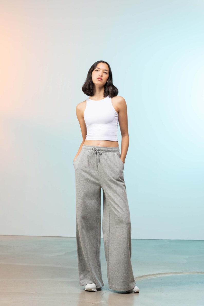 May include: A woman wearing a white tank top and grey wide-leg trousers. She is standing in front of a light blue background.