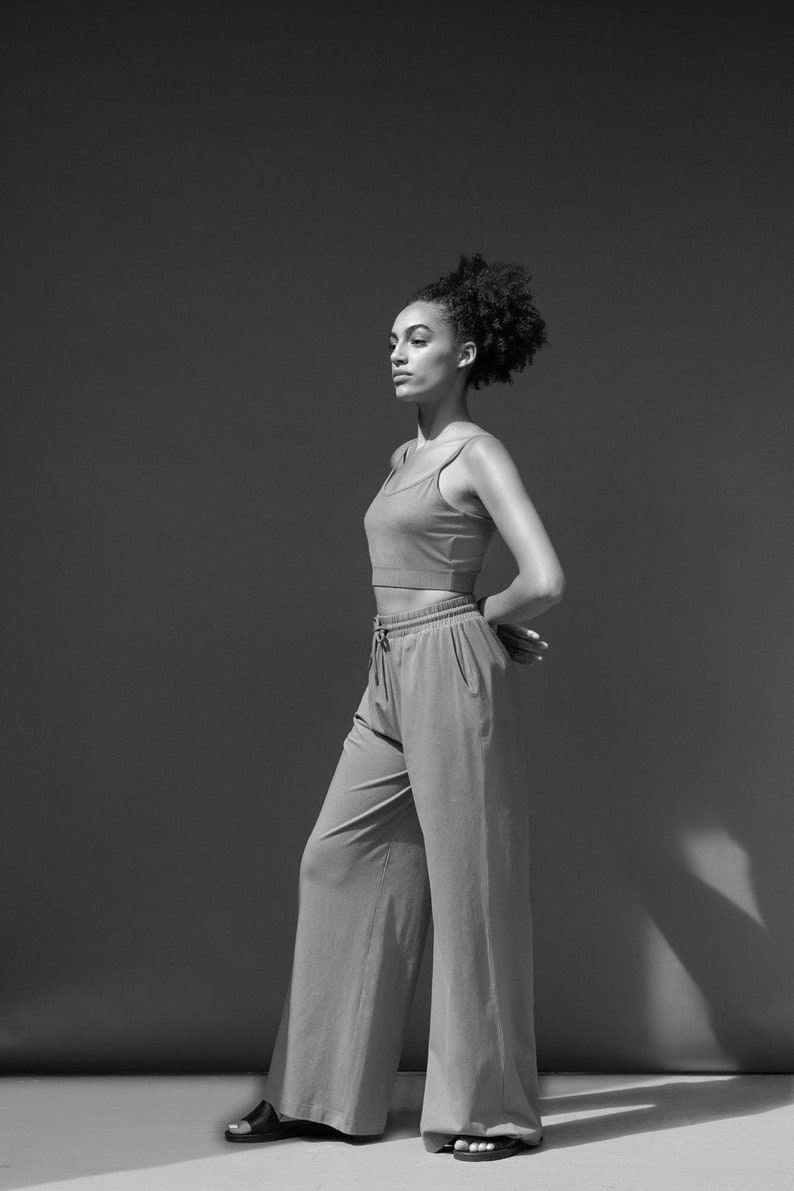May include: A woman wearing a black and white outfit. She is wearing a black tank top and white wide-leg trousers. She is standing in front of a grey background.