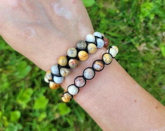 May include: Two beaded bracelets with natural stone beads in shades of yellow, brown, gray, and white. The beads are strung on black cords. The bracelets are worn on a wrist against a background of green grass.