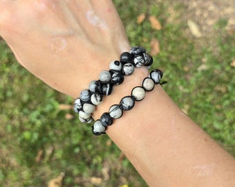 May include: Two beaded bracelets with black and white patterned beads. The bracelets are made of small, round beads strung together on a black cord. The beads have a mix of black, white, and gray colors. The bracelets are worn on a wrist.