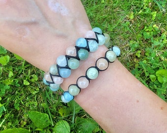 May include: Two beaded bracelets with a variety of pastel-colored stones, including light blue, pink, and green. The beads are strung on black cord. The bracelets are displayed on a person's wrist against a backdrop of green grass.