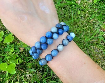 May include: Two beaded bracelets with blue and light blue stones. The bracelets are made with a black cord and are worn on a person's wrist. The background is green grass.