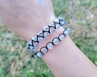 May include: Two clear quartz crystal bead bracelets with black string are displayed on a wrist. The larger bracelet has a woven design, while the smaller one has a simple string design. The background is blurred green grass.