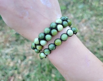 May include: Two jade bead bracelets. Each bracelet features round, light green beads strung on black cord. The bracelets are stacked on top of each other. The background is blurred green.