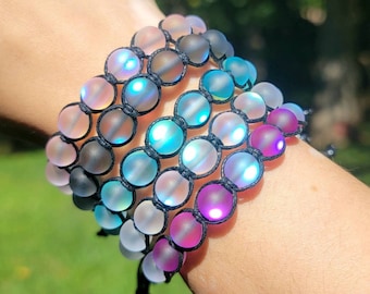 May include: Several bracelets with iridescent, frosted glass beads in shades of pink, blue, and purple are displayed. The beads are strung on black cord, creating a layered effect. The bracelets are on a wrist.