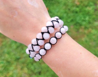 May include: Two rose quartz beaded bracelets. Each bracelet features light pink, round beads strung on black cord. The bracelets are worn together, showcasing the delicate color and texture of the gemstones.
