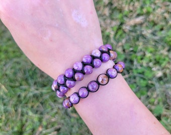 May include: Two beaded bracelets with purple and brown stones are displayed on a wrist. The bracelets are made with round, polished stones and black cord. The background is blurred, showing green grass.