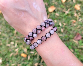 May include: Two beaded bracelets with pink and purple stones are displayed. The bracelets are strung on black cord. The larger bracelet has a woven design, while the smaller one has a simple string of beads. The background is blurred green grass.