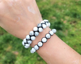 May include: Two white and gray beaded bracelets with black string are worn on a wrist. The bracelets are made of round beads. The background is a blurred green.