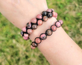 May include: Two bracelets with pink and black gemstone beads. The beads are strung on black cord. The bracelets are worn on a person's wrist. The background is blurred green grass.