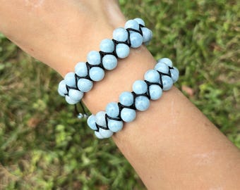 May include: Two aquamarine beaded bracelets. Each bracelet features two rows of light blue, round beads, connected by black cord. The bracelets are worn on a wrist, with a blurred green background.