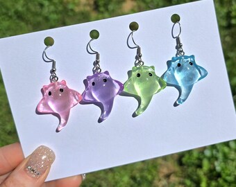 May include: Four pairs of colorful stingray earrings. Each earring features a translucent stingray design in pink, purple, green, and blue. The earrings have silver-colored hooks and small green accents at the top, displayed on a white card.