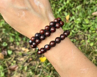 May include: Two beaded bracelets with dark red-brown stones are worn on a wrist. The bracelets are made with small, round beads strung on black cord. The background is blurred green grass.