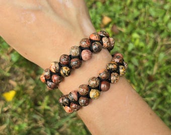 May include: Two double-strand beaded bracelets. The bracelets feature round, speckled beads in shades of brown, orange, and yellow, strung on black cord. The bracelets are worn on a wrist, with a blurred green background.