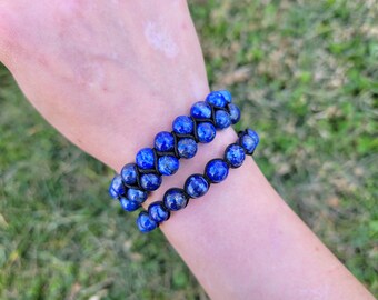May include: Two blue beaded bracelets. The bracelets are made of round, dark blue beads strung on black cord. One bracelet is a double strand, and the other is a single strand. The bracelets are displayed on a person's arm.