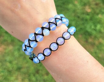 May include: Two beaded bracelets with opalescent beads and black string. The top bracelet has two rows of beads, while the bottom bracelet has one row. The bracelets are worn on a wrist against a green background.