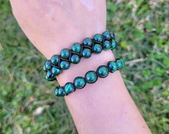 May include: Two green beaded bracelets. One bracelet has two rows of beads, and the other has one row. The beads are a dark green color with a reflective sheen. The bracelets are on a black cord.