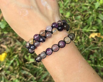 May include: Two beaded bracelets with purple, yellow, and clear stones. The beads are strung on black cord. The bracelets are worn on a wrist against a background of green grass.
