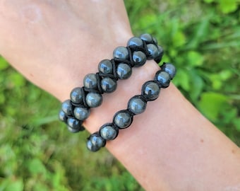 May include: Two beaded bracelets with dark gray, spherical beads are shown. The beads are strung on black cord, with a woven design. The bracelets are worn on a wrist, with green foliage in the background.