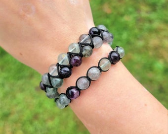 May include: Two beaded bracelets with a mix of green, purple, and clear fluorite stones. The beads are strung on black cord, creating a woven pattern. The bracelets are worn on a wrist, showcasing the natural stone jewelry.
