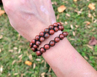 May include: Two red jasper beaded bracelets. The bracelets are made with round, reddish-brown beads and black cord. The bracelets are worn on a wrist, with the background showing green grass and fallen leaves.