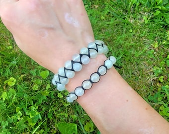 May include: Two beaded bracelets with light green beads and black string. The top bracelet has a woven design, while the bottom bracelet has a simple string design. The bracelets are displayed on a person's arm against a backdrop of green grass.