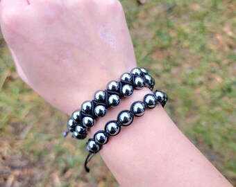 May include: Two bracelets with shiny, dark gray beads are worn on a wrist. The top bracelet has a woven design, while the bottom one is a single strand. Both are strung on black cord.