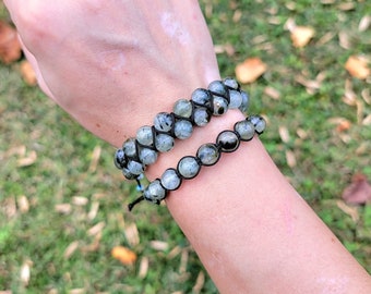 May include: Two beaded bracelets with light green and black speckled stones. The bracelets are made with black cord and are worn on a wrist against a blurred green background. One bracelet is double-stranded.