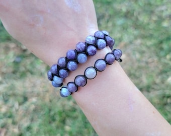 May include: Two beaded bracelets with purple and gray stones are worn on a wrist. The bracelets are made with black string. The background is blurred green grass.