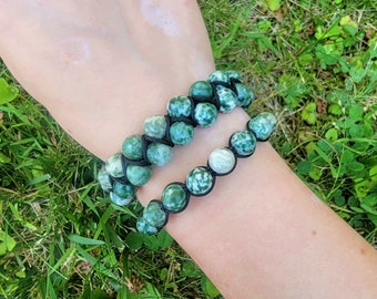 May include: Two beaded bracelets. The larger bracelet features two rows of green and white speckled beads, while the smaller bracelet has a single row. Both bracelets are strung on black cord. The bracelets are displayed on a person's arm against a backdrop of green grass.