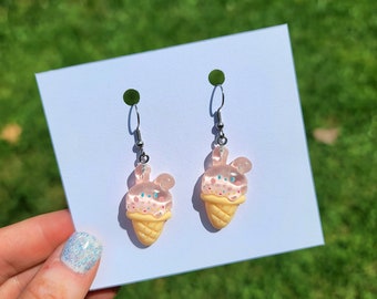 May include: A pair of dangle earrings featuring a light pink ice cream cone design with colorful sprinkles. The earrings have a clear, translucent appearance with silver-colored hooks. The ice cream cones have a bunny ear shape.