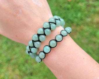 May include: Two jade green beaded bracelets. The bracelets are made with round, polished beads strung on black cord. One bracelet has a woven design, while the other is a single strand. The bracelets are shown on a person's wrist.