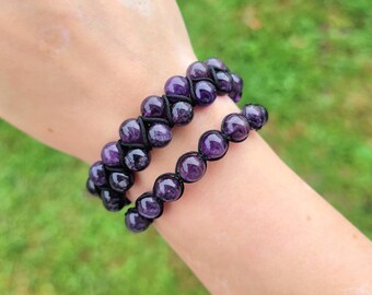 May include: Two amethyst beaded bracelets. The bracelets feature dark purple, round beads strung on black cord. The bracelets are worn on a wrist, with a blurred green background.