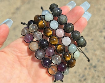 May include: A beaded bracelet featuring a variety of colorful gemstones, including tiger's eye, amethyst, and rose quartz. The beads are strung together with black cord, creating a unique and eye-catching accessory. The bracelet is held in a hand.