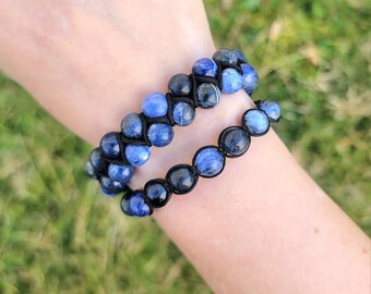 May include: Two beaded bracelets with blue and black stones are displayed on a wrist. The bracelets are made with a black cord, and the stones have a polished appearance. The background is blurred, with green grass visible.