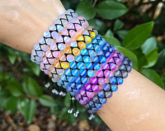 May include: A collection of colorful beaded bracelets. Each bracelet features iridescent, round beads in shades of pink, yellow, blue, and purple, strung on black cord. The bracelets are stacked on a wrist, showcasing their varying colors and textures.