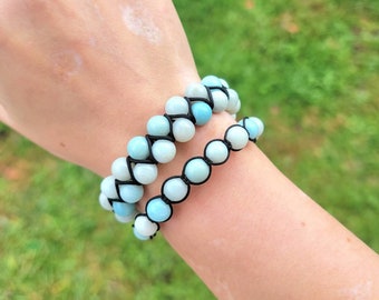 May include: Two beaded bracelets. The bracelets feature light blue and white beads strung on black cord. The bracelets are worn on a wrist, with a green grassy background.
