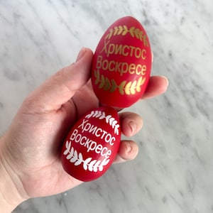 May include: Two red Easter eggs with gold and white lettering and leaf designs. The text on the eggs reads "Christ is Risen". The eggs are held in a hand against a white marble background.