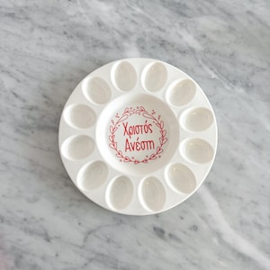 May include: A white ceramic egg serving platter with 12 oval indentations around the edge and a central well. The centre features red Greek text within a decorative wreath. The platter is on a marble surface.
