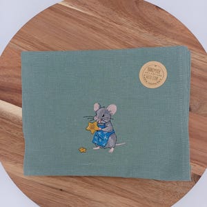 Könnte beinhalten: Ein gefaltetes, teal-farbenes Stoffserviette mit einer gestickten Maus, die eine blaue Schürze trägt und einen goldenen Stern hält. Ein kleinerer goldener Stern ist darunter gestickt. Ein runder Aufkleber mit dem Text "HANDMADE WITH LOVE" befindet sich auf der Serviette.