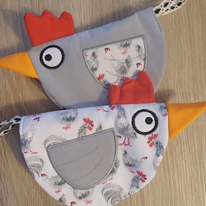 Könnte beinhalten: Zwei graue und weiße Stoff-Topflappen in Form von Hühnern mit roten Kämmen und orangefarbenen Schnäbeln. Die Topflappen haben einen weißen Hintergrund mit einem schwarz-weißen Hühnermuster.