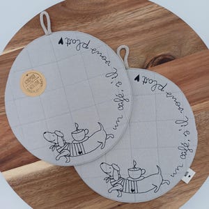 Könnte beinhalten: Zwei runde, hellgraue, gesteppte Topflappen mit einem Dackel-Design. Die Hunde tragen Pullover und stehen neben einer Kaffeetasse. Der Text "Il vous plaît un café?" ist am Rand gestickt. Jeder Topflappen hat eine Schlaufe zum Aufhängen.